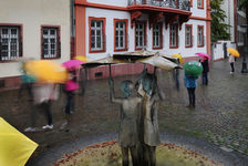Bildergalerie Brunnenanlagen Menschen mit Schirmen rund um den Ballplatzbrunnen Tag und Nacht und bei jedem Wetter stehen die drei lebensgroßen Mainzerinnnen aus Bronze am Ballplatz.